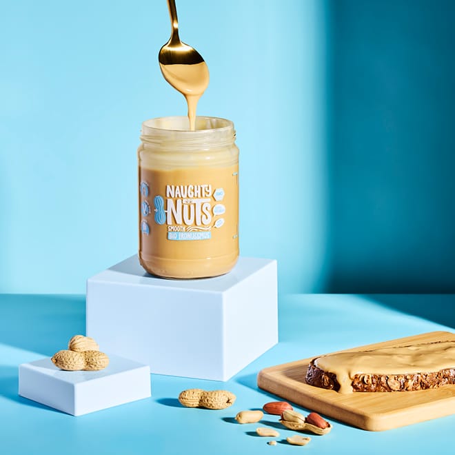 Bio Erdnussmus Smooth im Glas mit Löffel, Erdnüssen und Kuchen auf blauem Hintergrund