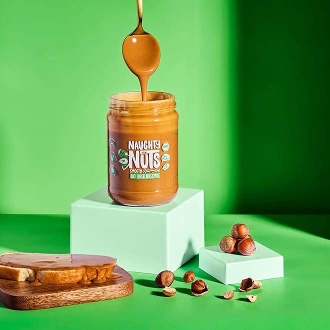 Löffel taucht in ein Glas Bio Haselnussmus Smooth, daneben Haselnüsse und ein Toast mit Mus