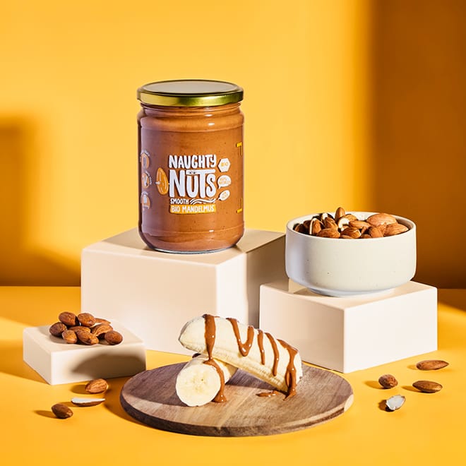 Bio Mandelmus Smooth (500g Glas) mit Banane und Mandeln auf gelbem Hintergrund