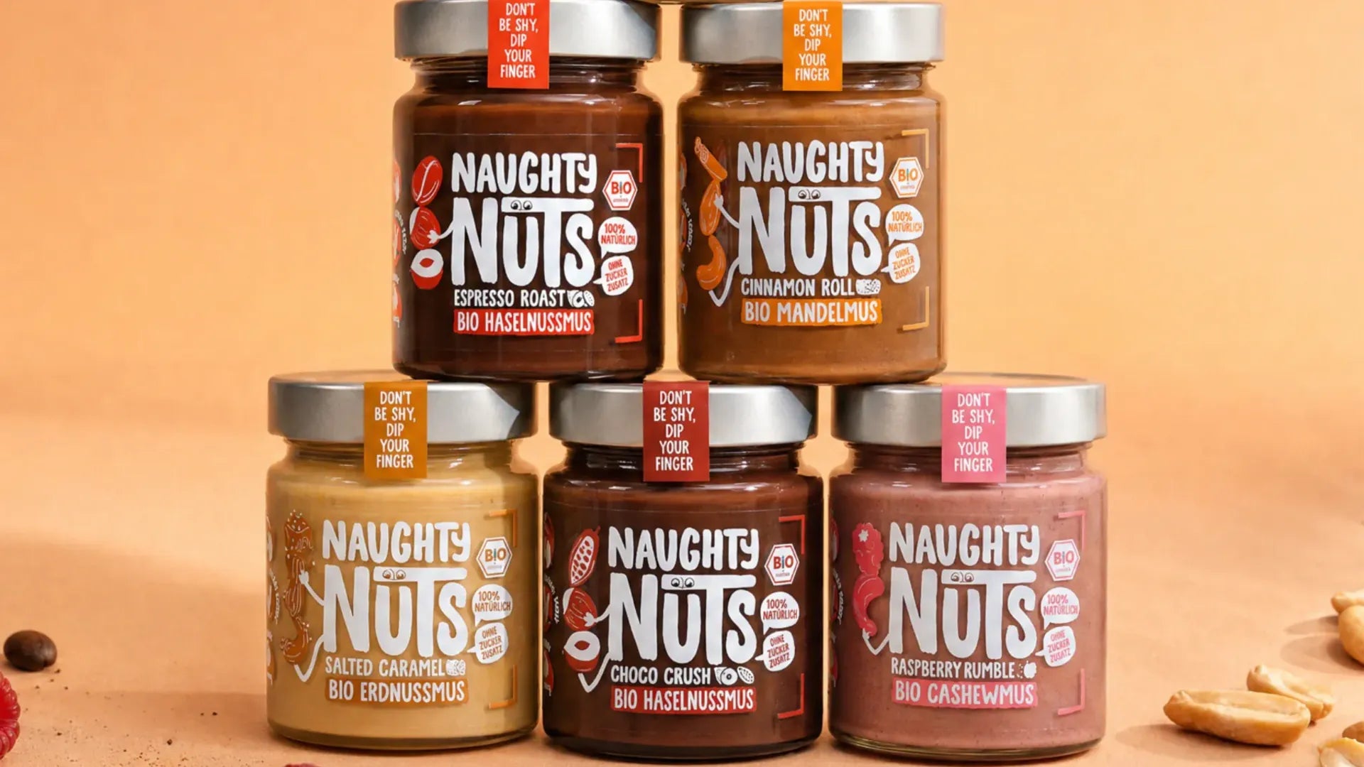 Fünf Gläser Naughty Nuts Bio Nussmus in verschiedenen Sorten gestapelt auf beigem Hintergrund